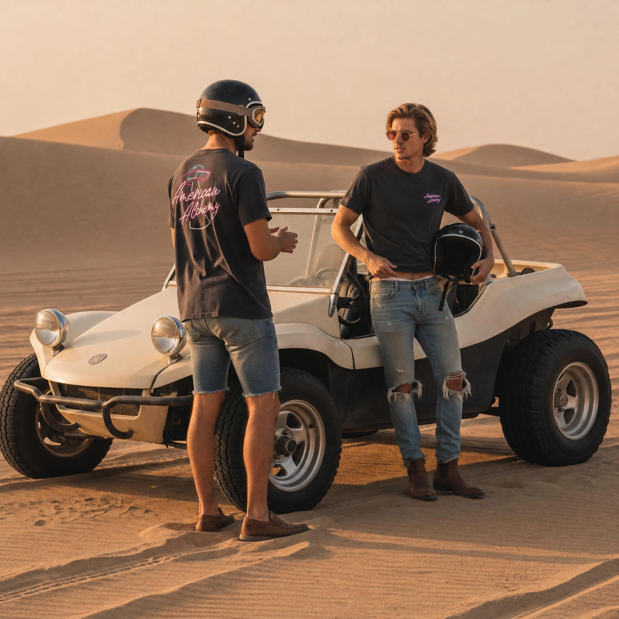 Two models by a dune buggy in the desert at golden hour, one wearing the black American Alchemy logo tee